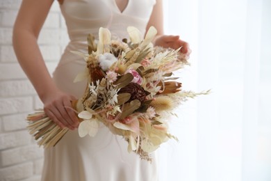 Bride holding beautiful dried flower bouquet near window at home, closeup