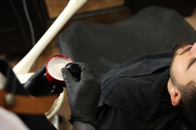 Professional hairdresser with shaving foam near bearded client in barbershop, closeup
