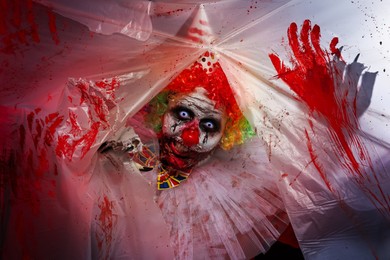 Terrifying clown staring through hole in torn bloodstained plastic film. Halloween party costume