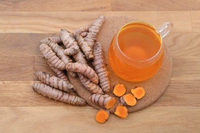 Photo of Glass cup of hot tea and fresh turmeric roots on wooden table, above view