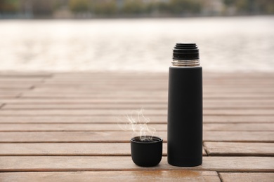 Black thermos and cap on wooden pier near river. Space for text