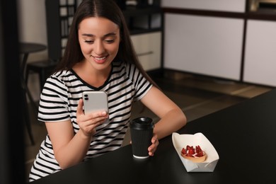 Happy young woman with paper cup of coffee and smartphone at table in hostel dining room