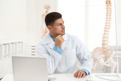 Male orthopedist with laptop near human spine model in office
