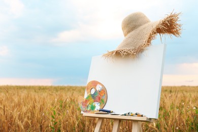 Wooden easel with blank canvas, painting equipment and hat in field. Space for text