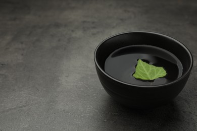 Photo of Black bowl with water and green leaf on grey table. Space for text
