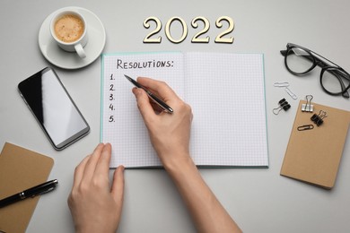 Woman filling list of resolutions for 2022 new year in notebook on light background, top view