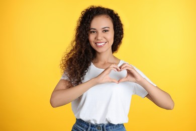 Happy young African-American woman making heart with hands on yellow background