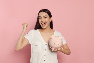 Emotional young woman with ceramic piggy bank on pale pink background