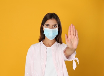 Woman in protective mask showing stop gesture on yellow background. Prevent spreading of coronavirus