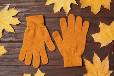 Stylish orange woolen gloves and dry leaves on wooden table, flat lay
