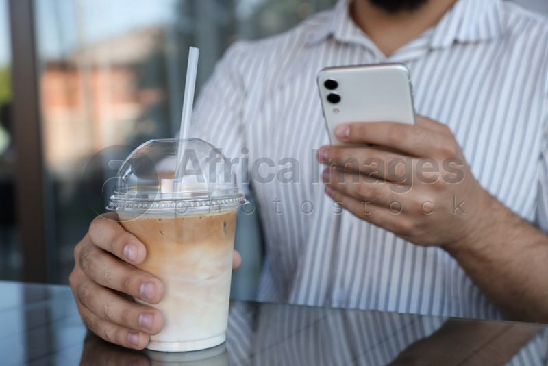 Man with smartphone and plastic takeaway cup of delicious iced coffee at table in outdoor cafe, closeup