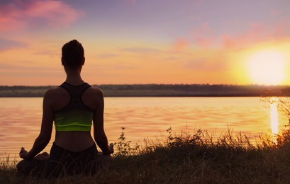 Woman meditating near river at sunset, back view. Practicing yoga