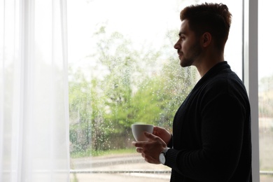 Thoughtful handsome man with cup of coffee near window indoors on rainy day