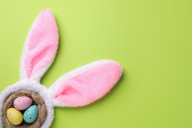 Headband with Easter bunny ears and dyed eggs in nest on green background, flat lay. Space for text