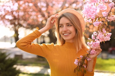 Young woman wearing stylish outfit near blossoming sakura in park. Fashionable spring look