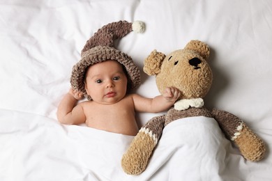 Cute little baby with toy bear lying under blanket in bed, top view