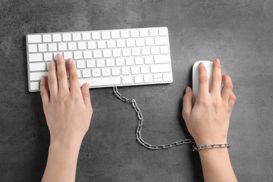 Woman chained to computer keyboard at grey table, top view. Loneliness concept