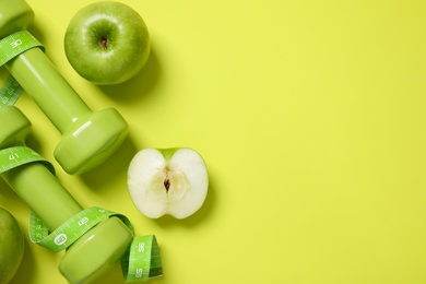 Fresh apples, measuring tape and dumbbells on green background, flat lay. Space for text