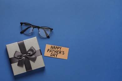 Card with phrase HAPPY FATHER'S DAY, eyeglasses and gift box on blue background, flat lay. Space for text