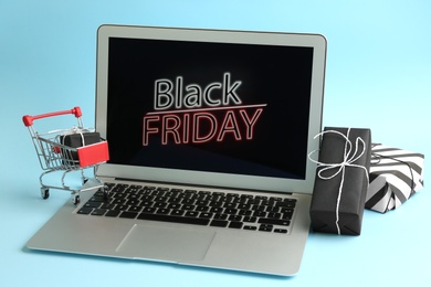 Laptop with Black Friday announcement, small shopping cart and gifts on light blue background