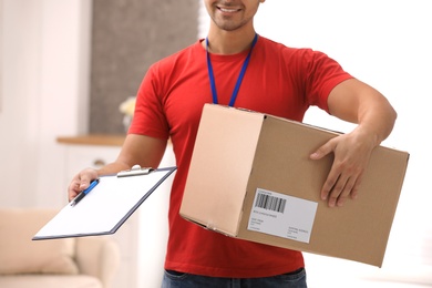 Young courier holding parcel and clipboard indoors, closeup. Delivery service
