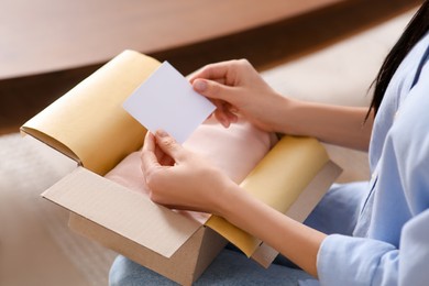Photo of Woman holding blank greeting card near package with gift indoors, closeup