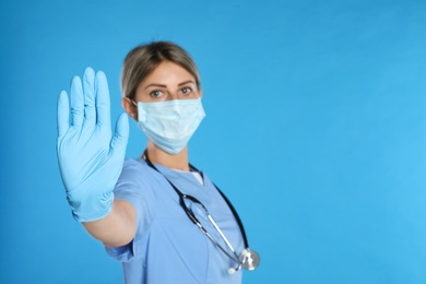 Doctor in protective mask showing stop gesture on light blue background, space for text. Prevent spreading of coronavirus