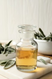 Bottle of essential sage oil and leaves on white wooden table, closeup