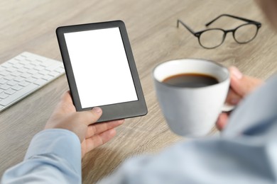 Man with cup of coffee using e-book reader at wooden table, closeup