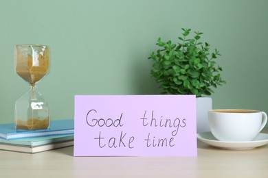 Phrase Good Things Take Time, coffee, houseplant and hourglass on table against light green background. Motivational quote