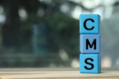 Photo of Abbreviation CMS of blue cubes on wooden table against blurred background, space for text. Content management system
