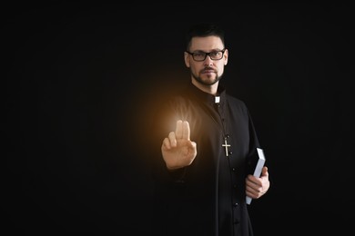 Priest with Bible making blessing gesture and holy light on dark background, space for text