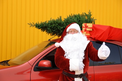 Photo of Authentic Santa Claus near car with fir tree and bag full of presents on roof against yellow background