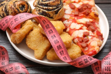 Photo of Plate of different unhealthy food and measuring tape on wooden table, closeup. Weight loss concept