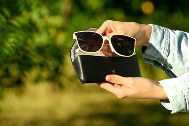 Photo of Woman holding sunglasses and case outdoors on sunny day, closeup. Space for text