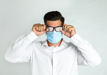Man wiping foggy glasses caused by wearing medical mask on light background