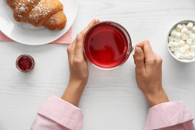 Woman holding glass cup of raspberry tea at white wooden table, top view. Delicious morning meal