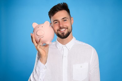 Photo of Happy young man with piggy bank on light blue background