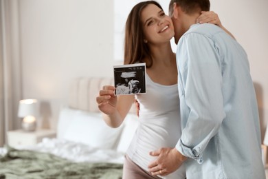 Photo of Young pregnant woman with her husband in bedroom, focus on ultrasound picture of baby