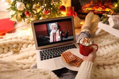 MYKOLAIV, UKRAINE - DECEMBER 23, 2020: Woman with sweet drink watching The Christmas Chronicles movie on laptop near fireplace at home, closeup. Cozy winter holidays atmosphere