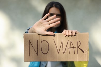 Photo of Young woman holding poster with words No War and showing stop gesture near light wall, focus on hand