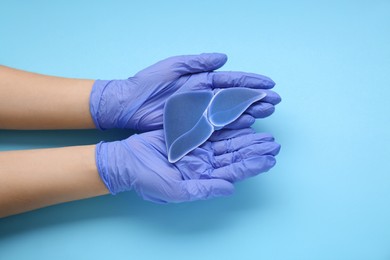 Photo of Doctor in gloves holding paper liver on light blue background, closeup and top view. Hepatitis treatment