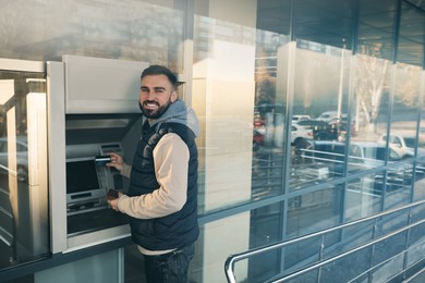 Photo of Young man using cash machine for money withdrawal outdoors