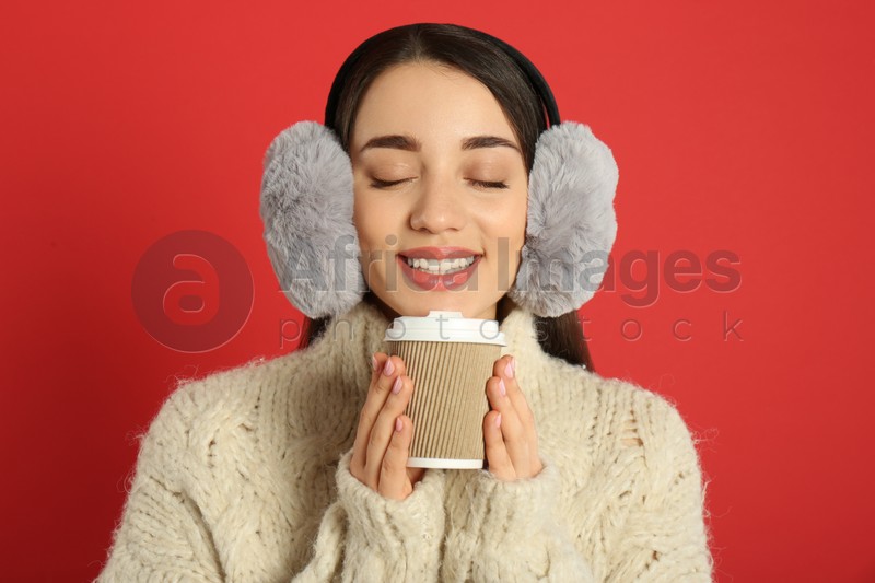 Beautiful young woman in earmuffs with cup of drink on red background