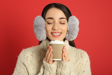 Beautiful young woman in earmuffs with cup of drink on red background