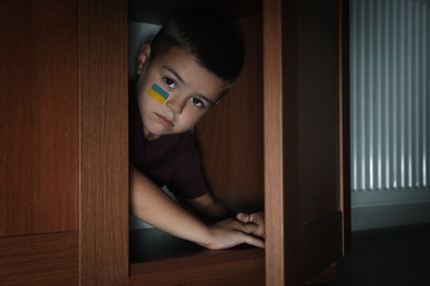 Sad little boy with picture of Ukrainian flag on cheek hiding in wardrobe. Stop war in Ukraine