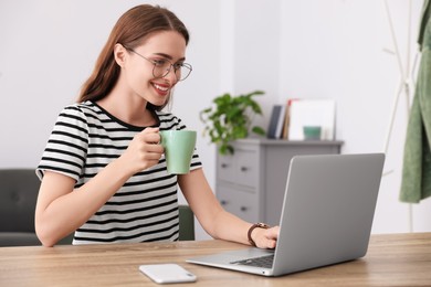Photo of Happy young woman with laptop and drink at table indoors