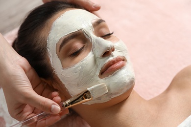 Cosmetologist applying white mask onto woman's face in spa salon, closeup