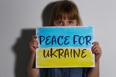 Sad little girl holding paper with phrase Peace for Ukraine near white wall