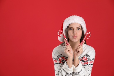 Pretty woman in Santa hat and Christmas sweater holding candy canes on red background, space for text